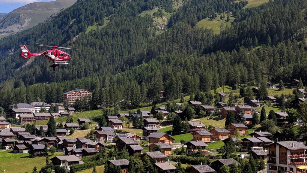 Hélicoptère rouge survolant un village alpin boisé en montagne.
